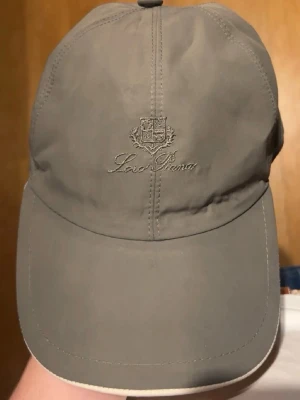 Grön loro piana keps - Snygg grön keps från Loro Piana med broderad logga framtill och klassisk böjd skärm. Insidan har ett rutigt foder och etikett med 'Storm System'. Perfekt accessoar för en stilren look.