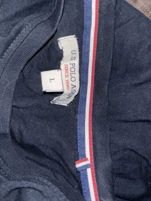Mörkblå t-shirt från U.S. Polo Assn - Snygg mörkblå t-shirt från U.S. Polo Assn med röd broderad logga på bröstet och en randig detalj i rött, vitt och blått på insidan av kragen. Tillverkad i mjuk bomull för skön känsla hela dagen.