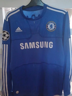 Chelsea matchtröja Adidas Frank Lampard  - Blå långärmad Chelsea FC matchtröja från Adidas med Champions League-märke på ärmen och Samsung som sponsor på bröstet. Tröjan har vita detaljer och klubbens emblem på bröstet. Materialet är lätt och ventilerande mesh, perfekt för fotboll. Storlek M,  tröjan är äkta.