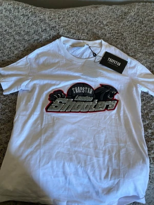Vit Trapstar t-shirt med Shooter-tryck - Säljer en vit t-shirt från Trapstar med stort broderat Shooter-motiv på bröstet i svart, grått och rött. Klassisk rund hals och korta ärmar. Perfekt för dig som vill ha en cool och unik streetwear-look.