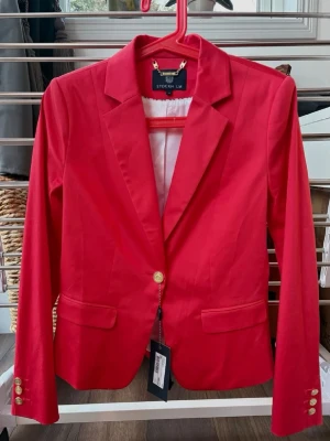 Röd kavaj från STOCKH LM - Stilren röd kavaj från STOCKH LM med klassisk krage, guldknappar och två fickor framtill. Figursydd modell med snygg passform och helfodrad insida. Perfekt för att lyfta din outfit med en färgklick. Aldrig använd, prislapp finns kvar! 