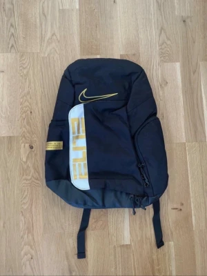 Svart Nike Elite ryggsäck - Snygg svart ryggsäck från Nike med gula detaljer och texten ELITE på framsidan. Ryggsäcken har flera fack, justerbara axelband med vaddering och en stor Nike-logga. Perfekt för skolan eller träningen. Tillverkad i slitstarkt syntetmaterial. Ny med prislapp!