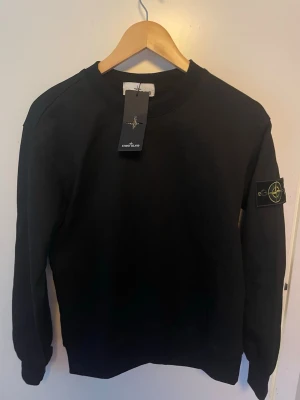 Svart sweatshirt från Stone Island - Cool svart sweatshirt från Stone Island med klassisk rund hals och lång ärm. Ikonisk logotyp-patch på vänster ärm som fästs med knappar. Tröjan är tillverkad i mjukt bomullsmaterial och har en stilren design som passar perfekt till jeans eller joggers.