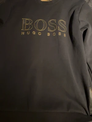 Svart sweatshirt från Hugo Boss - Snygg svart sweatshirt från Hugo Boss med stort BOSS-tryck i guld på bröstet. Tröjan har rund halsringning och ribbade muddar. Materialet är mjukt och bekvämt, perfekt för en stilren look. Diskret BOSS-detalj på insidan av kragen.