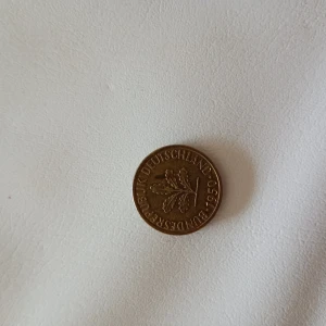 Tysk 5 Pfennig mynt 1950 - Säljer ett tyskt 5 Pfennig mynt från 1950. Myntet är i bronsfärg och har en ekkvist på ena sidan och siffran 5 med texten 'PFENNIG' på andra sidan. Perfekt för samlare av utländska mynt eller historiska föremål.