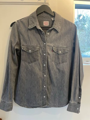 Grå jeansskjorta från Levi's - Snygg grå jeansskjorta från Levi's med klassisk krage, två bröstfickor med knappar och vita tryckknappar längs hela framsidan. Skjortan har lång ärm och är tillverkad i slitstark bomull. Perfekt för en avslappnad och cool stil.