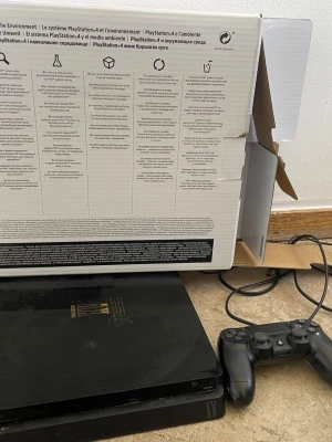 PlayStation 4 Slim Final Fantasy XV - PlayStation 4 Slim med specialdesign för Final Fantasy XV. Konsolen är svart med guldfärgad logotyp och text på ovansidan. Medföljer en svart DualShock 4-kontroll och strömkabel. Perfekt för dig som älskar gaming och Final Fantasy.