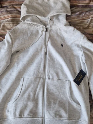 Ljusgrå hoodie från Polo Ralph Lauren - (Säljer den pga det inte är min stil) (Helt ny hoodie / zip up, pris kan diskuteras! )    snygg ljusgrå hoodie från Polo Ralph Lauren med dragkedja, två stora fickor framtill och klassisk mörkblå logga broderad på bröstet. Hoodien har justerbar huva med snören och är tillverkad i mjukt bomullsmaterial. Perfekt för en chill och stilren look.