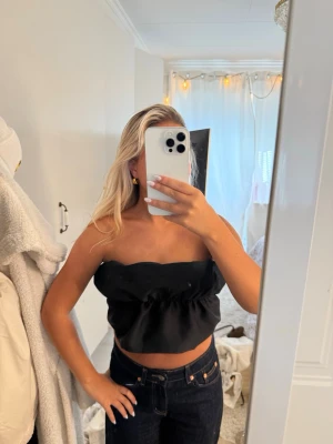 Zara Svart bandeau topp med volang - Trendig svart bandeau topp med volangdetalj runt om. Axelbandslös och croppad modell som ger en snygg siluett. Perfekt att matcha med höga jeans eller kjol för en cool look.