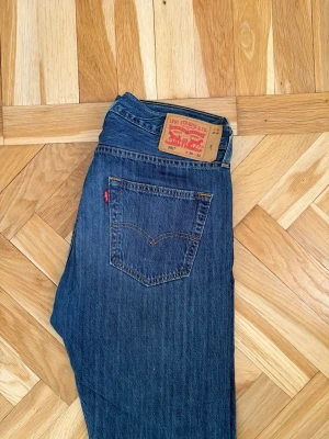Levi's 501 blå raka jeans - Klassiska blå Levi's 501 jeans med rak passform och fem fickor. Jeansen har den ikoniska röda Levi's-lappen på bakfickan och patch i läder bak i midjan. Tillverkade i slitstark denim med knappgylf. Perfekta för en avslappnad och tidlös look.