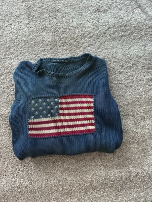 Ralph Lauren stickad tröja - Mörkblå stickad tröja från Ralph Lauren med en stor amerikansk flagga i rött, vitt och blått på bröstet. Tröjan har rund halsringning och ribbstickade muddar vid ärmslut och nederkant. Perfekt för dig som vill ha en klassisk och stilren look. Köpt på Vinted men var för liten 