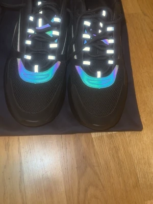 Svarta sneakers med reflexdetaljer - Säljer ett par svarta sneakers med reflekterande detaljer och meshpaneler. Skorna har en chunky sula och inslag av iriserande färg på ovandelen. Klassisk snörning och logga på plösen. Perfekta för dig som vill sticka ut med stilrena men unika sneakers. Det bara ställa frågor! Pris kan diskuteras😁