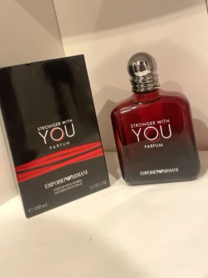 Stronger With You Parfum 100ml Armani - Stronger With You Parfum från Emporio Armani i en elegant mörkröd glasflaska med silverfärgad rund kork. Parfymen har en modern och stilren design med vita och röda detaljer på både flaska och kartong. Perfekt för dig som gillar exklusiva dofter.