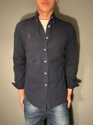 Mörkblå skjorta från Polo Ralph Lauren - Säljer en mörkblå skjorta från Polo Ralph Lauren med klassisk knappslå och vit knappdetalj. Skjortan har lång ärm och ikonisk röd logobrodyr på bröstet. Perfekt för dig som gillar stilrena plagg med premiumkänsla.
