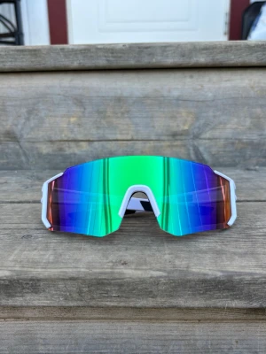 Sportiga spegelglasade solglasögon - Säljer ett par riktigt snygga sportiga solglasögon med spegelglas i grönt, blått och lila. De har en vit ram och en futuristisk, kantig form som täcker bra. Perfekta för dig som vill sticka ut och skydda ögonen mot solen.