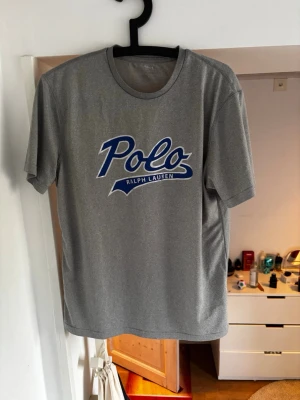 Grå t-shirt från Polo Ralph Lauren - Säljer en grå t-shirt från Polo Ralph Lauren med blå och vit logga på bröstet. T-shirten har rund hals och korta ärmar, perfekt för dig som gillar klassisk och stilren design. Materialet känns mjukt och bekvämt mot huden. Storlek S men passar som en storlek M