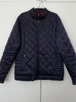 Mörkblå quiltad jacka från J&L - Quiltad mörkblå jacka från J&L med dragkedja framtill och två fickor med guldfärgade dragkedjor. Jackan har ståkrage med detalj och ett rutmönster över hela ytan. Perfekt för kyliga dagar och snygg att matcha med jeans.