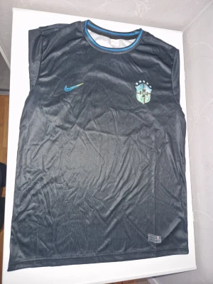 Svart Brasilien Nike fotbollströja - Cool svart fotbollströja från Nike med Brasilien-emblem på bröstet och blå detaljer vid halsen. Tröjan är ärmlös och tillverkad i ett lätt och ventilerande material som passar perfekt för träning eller match.