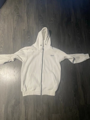 Vit hoodie från BOSS med dragkedja - Snygg vit hoodie från BOSS med dragkedja framtill och två stora fickor. Klassisk design med huva och ribbade muddar. Diskret BOSS-logga på bröstet. Perfekt för en clean och stilren look.