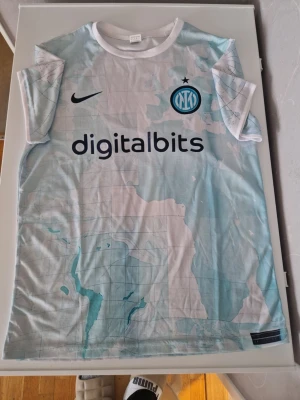 Inter Milan bortatröja 2022/23 - Snygg Inter Milan bortatröja från säsongen 2022/23 med ljusblått och vitt kartmönster, svart Nike-logga och klubbmärke på bröstet. Digitalbits och Lenovo som sponsorer. Tillverkad i lätt och ventilerande polyester, perfekt för fotboll eller träning.