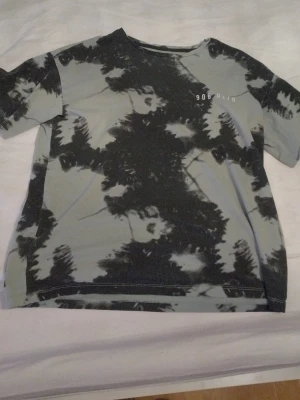 Svart och grå tie-dye t-shirt - Cool t-shirt med svart och grå tie-dye mönster och tryck med siffrorna '906.03/0' på bröstet. T-shirten har en klassisk passform och korta ärmar, perfekt för dig som gillar en avslappnad och trendig stil.