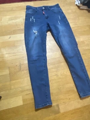 Blå  jeans med slitningar - Säljer ett par blå  jeans med coola slitningar och distressed detaljer på framsidan. Jeansen har klassisk femficksdesign och knappstängning. Perfekt för dig som gillar en trendig och avslappnad look.