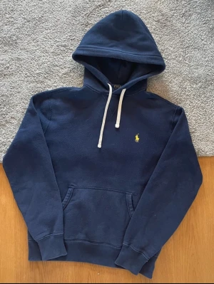 Mörkblå hoodie från Polo Ralph Lauren - Snygg mörkblå hoodie från Polo Ralph Lauren med klassisk huva och vita snören. Tröjan har en gul broderad logga på bröstet och en stor magficka. Perfekt för en avslappnad och cool look. Materialet är mjukt och bekvämt, troligtvis bomullsblandning.