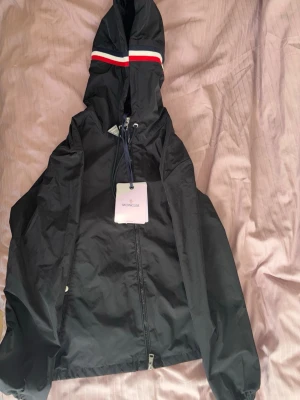 Svart vindjacka med huva från Moncler - Snygg svart vindjacka från Moncler med dragkedja framtill och justerbar huva. Huvan har en röd, vit och blå rand som ger en sportig vibe. Jackan är tillverkad i lätt och slitstarkt material, perfekt för blåsiga dagar.