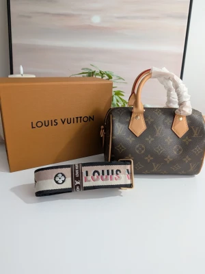 Iouis Vuitton Nano Speedy Bandoulière 20 - Detta är en kompakt handväska i bowlingmodell, utförd i ett ikoniskt brunt canvasmaterial. Ytan pryds av det välkända monogrammönstret i en ljusare beige ton, vilket skapar en tidlös kontrast mot den mörka bakgrunden.Väskan flankeras av en stor, klassisk saffransgul förvaringsbox med varumärkets namn tryckt i kontrasterande mörkblått. Den sportiga remmen ligger placerad framför väskan, vilket visar på möjligheten att variera stilen från elegant handburen till en modern crossover-look.