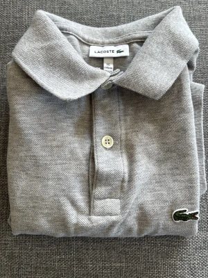 Grå pikétröja från Lacoste - Klassisk grå pikétröja från Lacoste med krage, knappar framtill och den ikoniska krokodilloggan broderad på bröstet. Perfekt basic till garderoben och enkel att matcha med olika stilar och storlek 176 cm.