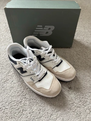 New Balance 550 vita och svarta sneakers - Snygga New Balance 550 sneakers i vitt och svart med beige detaljer. Skorna har klassisk låg siluett, läder- och mockadetaljer samt tydlig New Balance-logga på sidan.