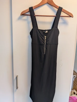 Svart klänning med dragkedja Divided - Snygg svart klänning från Divided med breda axelband och markerad v-ringning. Klänningen har en dekorativ dragkedja framtill och är figurnära i modellen. Perfekt för dig som gillar enkel men edgy stil.