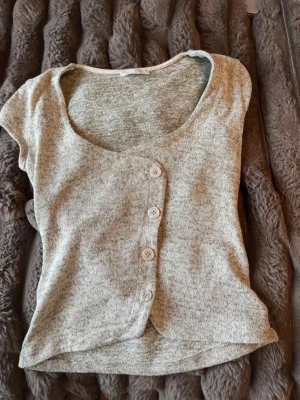 Grå ribbad topp med knappar - Grå ribbad topp med korta ärmar och snygg knappdetalj framtill. Toppen har en rundad halsringning och är tillverkad i ett mjukt, stretchigt material som sitter bekvämt. Perfekt att styla med jeans eller kjol.