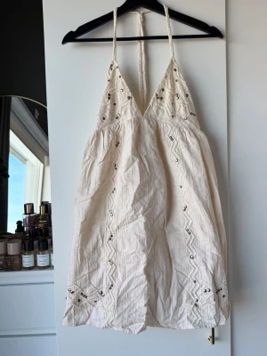 Vit kort klänning med paljettdetaljer - Säljer en vit kort klänning med tunna axelband och v-ringning. Klänningen har broderade mönster och små paljetter som ger extra glans. Tillverkad i ett lätt bomullstyg, perfekt för varma dagar. Snygg boho-vibe och avslappnad passform.