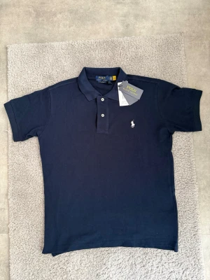 Marinblå pikétröja från Polo Ralph Lauren - Klassisk marinblå pikétröja från Polo Ralph Lauren med korta ärmar, krage och två knappar framtill. Tröjan har den ikoniska broderade loggan på bröstet och är tillverkad i mjuk bomull. Perfekt för en stilren och avslappnad look.