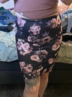 Svart blommig pennkjol med rosa toner - Snygg svart pennkjol med rosa och lila blommönster. Kjolen har en kroppsnära passform och går till knäna. Perfekt för dig som gillar feminina och trendiga plagg. Materialet känns mjukt och stretchigt, vilket gör den bekväm att bära hela dagen.