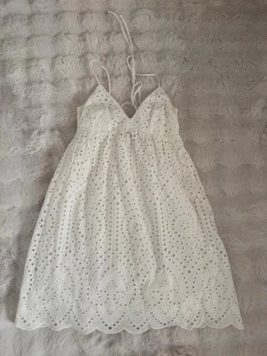 Vit somrig klänning med broderi - En klänning som är perfekt till sommaren eller till studenten. Det är en fin vit klänning med broderade detaljer och tunna axelband som knyts i nacken. Klänningen har v-ringning, öppen rygg och vackert hålmönster i tyget. Perfekt för varma dagar och har en romantisk vibe.
