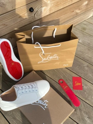 Vita Christian Louboutin sneakers - Snygga vita sneakers från Christian Louboutin med klassisk röd sula. Skorna har en stilren design med vita skosnören och detaljer i läder. Perfekta för dig som vill sticka ut med exklusiva och trendiga sneakers.