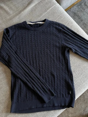 Mörkblå kabelstickad tröja Jack&Jones - Säljer en mörkblå kabelstickad tröja från J.Lindeberg. Tröjan har rund halsringning, långa ärmar och ribbade muddar. Klassisk och stilren design som passar perfekt till jeans eller chinos. Materialet känns mjukt och bekvämt.