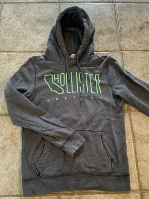 Mörkgrå Hollister hoodie - Mörkgrå hoodie från Hollister med limegrönt tryck på bröstet där det står 'Hollister Surfing' och en Kalifornien-siluett. Klassisk känguruficka och dragsko i huvan. Mjuk bomullsblandning, perfekt för chill dagar.