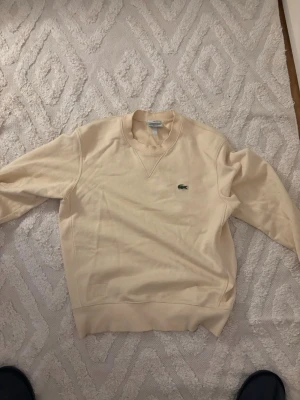 Beige sweatshirt från Lacoste - Snygg beige sweatshirt från Lacoste med klassisk krokodillogga på bröstet. Tröjan har rund halsringning, ribbade muddar och är i mjukt material. Perfekt för en avslappnad och stilren look.