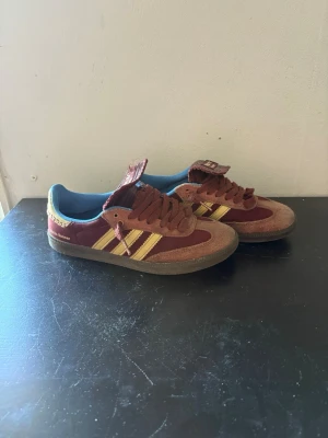 Adidas samba Wales bonner vinröd - Snygga Adidas samba Wales bonner i vinrött mocka och textil med gula ränder och blå insida. Klassisk retrostil med låg profil och gummisula. Perfekta för dig som gillar färgstarka och unika sneakers med old school-vibe.