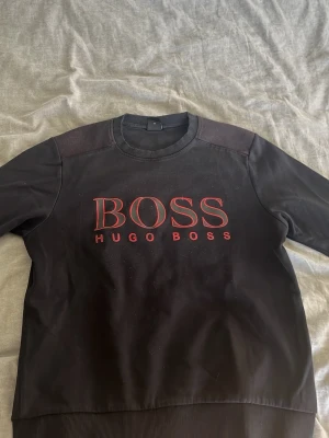 Svart sweatshirt från Hugo Boss - Svart sweatshirt med röd BOSS-logga och texten HUGO BOSS på bröstet. Tröjan har rund halsringning, ribbade muddar och är i slim fit-modell. Perfekt för en stilren och modern look.
