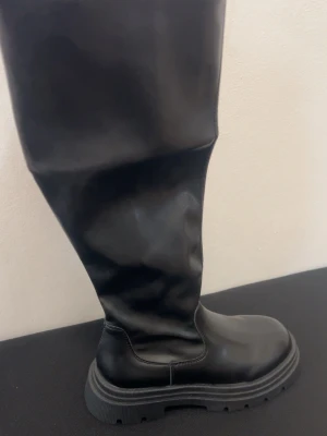Svarta höga chunky stövlar - Säljer ett par svarta höga stövlar med chunky sula och rund tå. Stövlarna är i ett glansigt material som ser ut som syntetläder och har en modern, stilren look. Perfekta för att lyfta vilken outfit som helst.