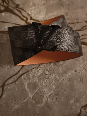 L V bälte svart - Snyggt L V bälte i svart och grått Damier-mönster med stor L V-logga i svart metall. Bältet är gjort i läder med ljusbrun insida och klassisk design. Perfekt accessoar för att lyfta din outfit.