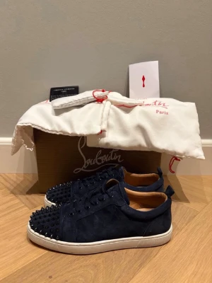 Christian Louboutin skor - Snygga mörkblå sneakers från Christian Louboutin i mocka med svarta nitar på tån och vita sulor. Skorna har klassisk låg modell och lyxig känsla med läderdetaljer invändigt. Perfekta för dig som vill sticka ut med en edgy och exklusiv stil.