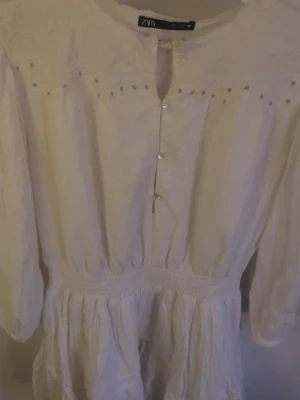 Vit klänning med broderi från ZARA - Söt vit klänning från ZARA med broderade detaljer upptill och knappar framtill. Klänningen har lång ärm, markerad midja med smock och volang i kjolen. Perfekt för en fräsch och romantisk look.