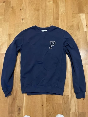 Mörkblå sweatshirt från Pier One - Mörkblå sweatshirt från Pier One med rund halsringning och ett broderat P på bröstet. Tröjan har långa ärmar och ribbade muddar vid ärmslut och nederkant. Perfekt för en avslappnad och stilren look.