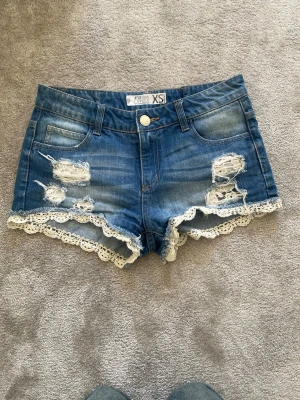 Blå denimshorts med spetskant - Säljer ett par blå denimshorts från FBsisters i storlek XS. De har slitna detaljer framtill och en vit spetskant längs bensluten som ger en söt touch. Klassisk femficksmodell med knapp och dragkedja. Perfekta för sommarens alla äventyr!