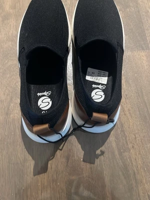 Svarta slip-on sneakers från Sprox - Svarta slip-on sneakers från Sprox med ovandel i mesh-tyg och vit, chunky sula. Skorna har bruna detaljer i läderimitation vid hälen och elastiska partier för enkel påtagning. Perfekta för en avslappnad och trendig stil.
